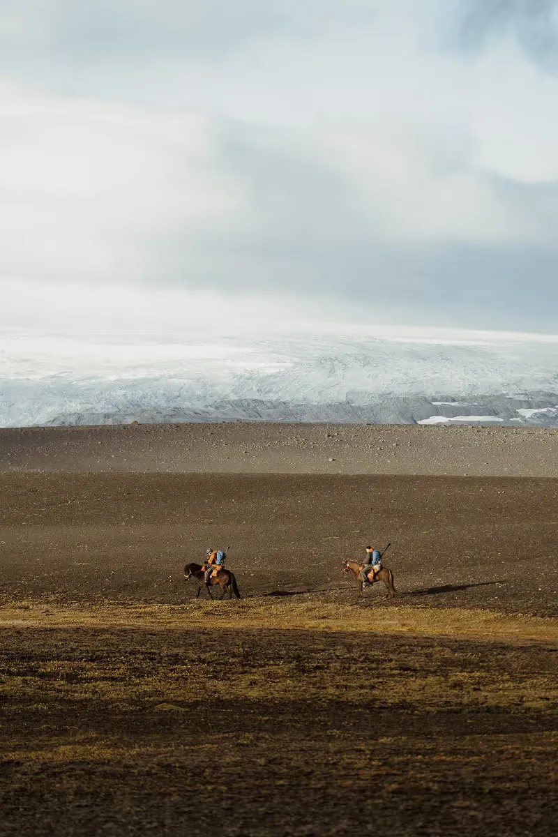 Two horseback riders crossing barren landscape with glacier in background
