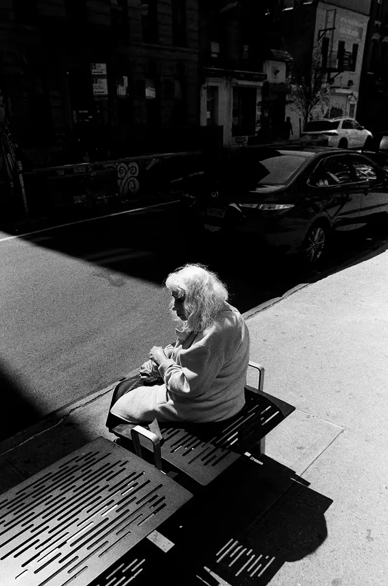 Elderly woman with white hair sitting on sunny sidewalk bench