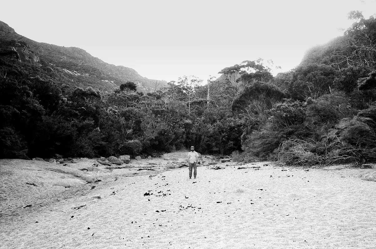 Lone figure standing on sandy riverbed surrounded by forested hills