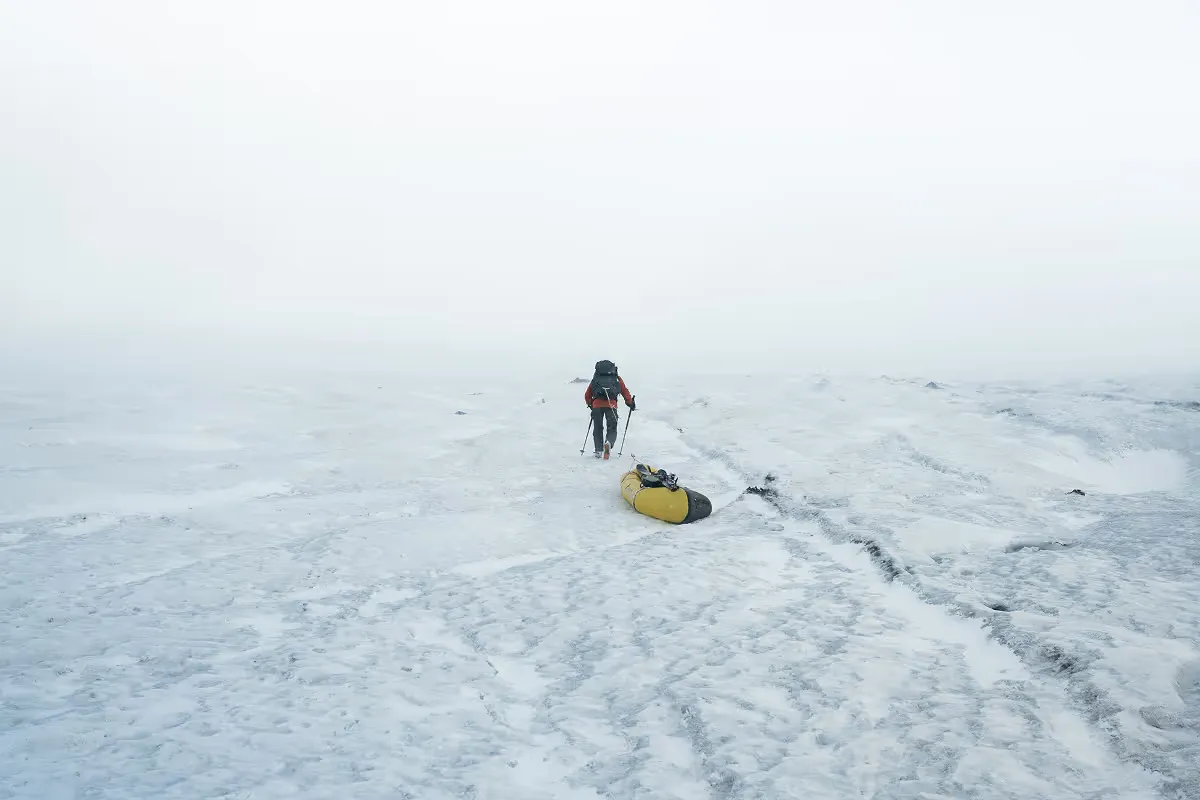 Solo hiker dragging yellow sled across vast snowy whiteout landscape