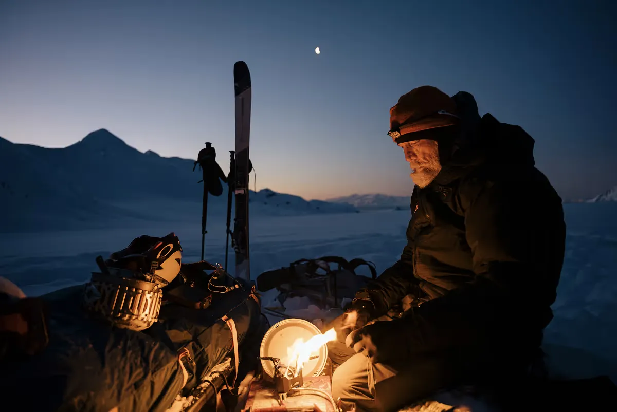 Geoff cooking with camp stove at twilight beside ski equipment